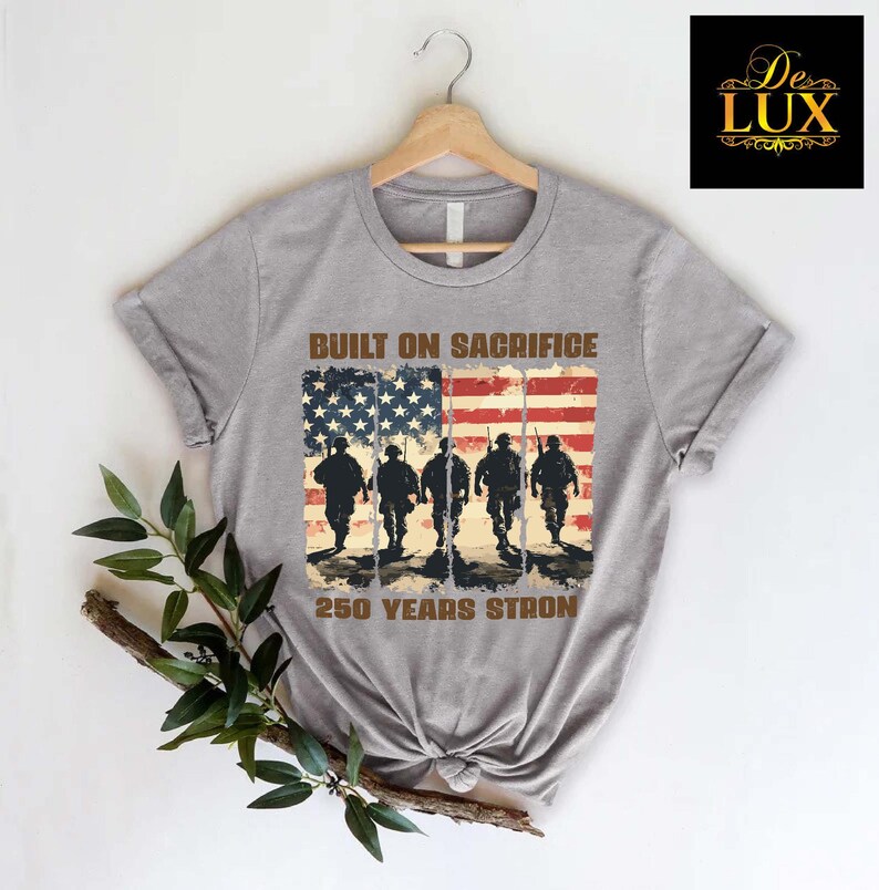 May include: A grey t-shirt with a distressed American flag design and soldier silhouettes. The text "BUILT ON SACRIFICE" and "250 YEARS STRONG" are printed above and below the flag graphic. A De Lux logo is in the upper right corner.