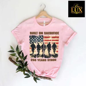 May include: Pale pink t-shirt featuring an American flag design with soldier silhouettes. The text reads "BUILT ON SACRIFICE" and "250 YEARS STRONG". The t-shirt is displayed on a wooden hanger. The design is patriotic.