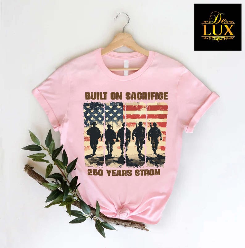 May include: Pale pink t-shirt featuring an American flag design with soldier silhouettes. The text reads "BUILT ON SACRIFICE" and "250 YEARS STRONG". The t-shirt is displayed on a wooden hanger. The design is patriotic.