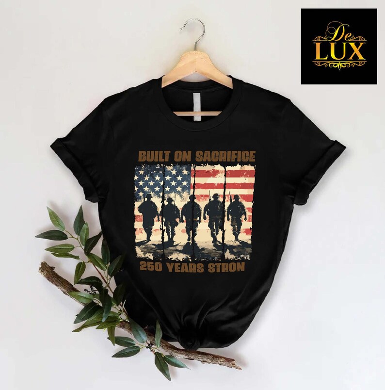 May include: Black t-shirt with a graphic of soldiers marching in front of an American flag. The text reads "BUILT ON SACRIFICE" above the flag and "250 YEARS STRON" below. The shirt is displayed on a wooden hanger.