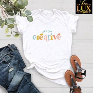 May include: A white short-sleeve t-shirt featuring the text let's get creative in a multi-colored, rounded font. The shirt is displayed alongside blue denim jeans and a pair of brown strappy sandals, with a leafy green branch visible.