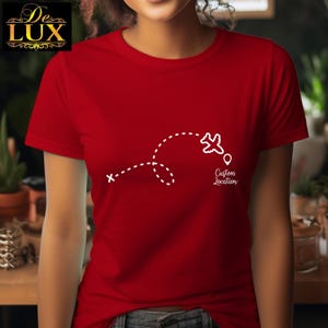 Pu&ograve; includere: Una maglietta rossa con una grafica bianca di un aereo che segue una linea tratteggiata, con la scritta "Custom Location" sotto un segnaposto. La maglietta ha maniche corte e girocollo.