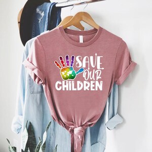 Peut inclure: T-shirt rose poussi&eacute;reux &agrave; manches retrouss&eacute;es. Le t-shirt pr&eacute;sente un motif d'empreinte de main color&eacute;e avec une carte du monde et le texte "SAVE OUR CHILDREN" en blanc. Le t-shirt est suspendu &agrave; un cintre en bois.