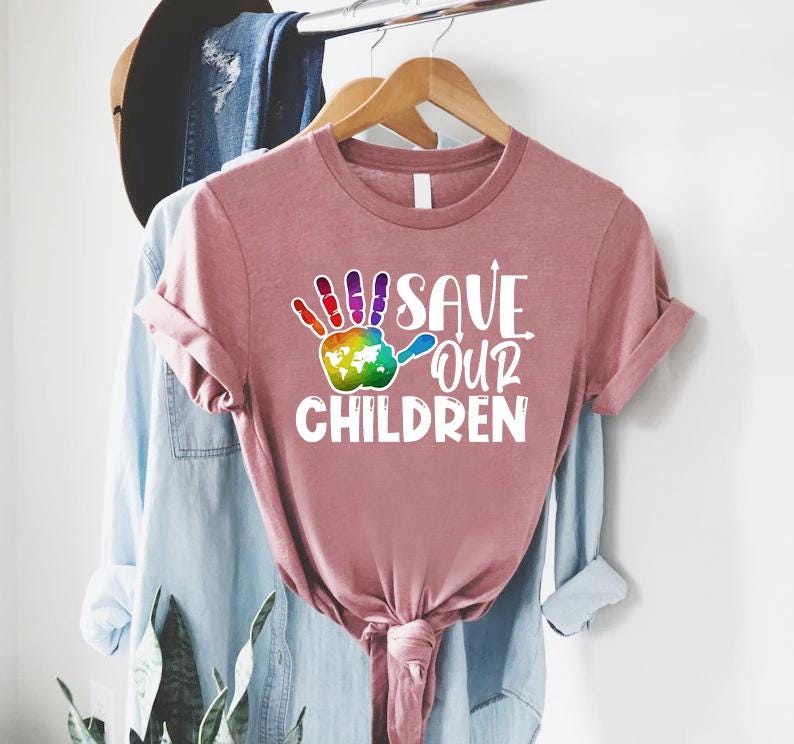 Peut inclure: T-shirt rose poussi&eacute;reux &agrave; manches retrouss&eacute;es. Le t-shirt pr&eacute;sente un motif d'empreinte de main color&eacute;e avec une carte du monde et le texte "SAVE OUR CHILDREN" en blanc. Le t-shirt est suspendu &agrave; un cintre en bois.