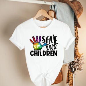 Peut inclure: T-shirt blanc avec un motif de main color&eacute;e et le texte "SAVE OUR CHILDREN" en noir. L'empreinte de main pr&eacute;sente un motif arc-en-ciel avec une carte du monde dans la paume. Le t-shirt est suspendu &agrave; un cintre en bois.