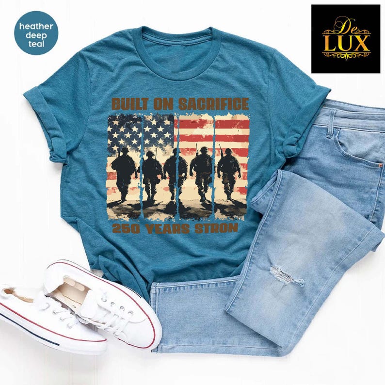May include: Heather deep teal t-shirt featuring the text "BUILT ON SACRIFICE" and "250 YEARS STRONG" above an American flag design with soldier silhouettes. The shirt is styled with light blue jeans and white trainers.