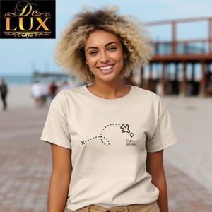 Pu&ograve; includere: Una maglietta beige con una linea tratteggiata nera, una grafica di un aereo e le parole "Custom Location". Il logo De Lux &egrave; nell'angolo in alto a sinistra. La maglietta &egrave; indossata da una persona con i capelli biondi ricci.