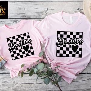 May include: Two light pink t-shirts with the word "Besties" in a black, cursive font, set within a black and white checkered square, and a black heart. One shirt is smaller than the other.
