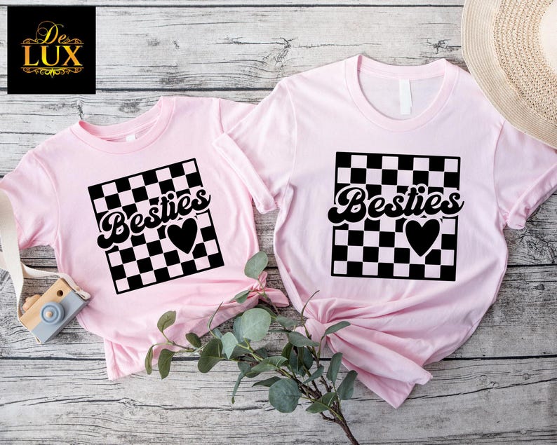 May include: Two light pink t-shirts with the word "Besties" in a black, cursive font, set within a black and white checkered square, and a black heart. One shirt is smaller than the other.