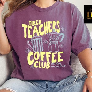 K&ouml;nnte beinhalten: Ein lila T-Shirt mit gelbem Aufdruck "Tired Teachers Coffee Club" mit Illustrationen einer Kaffeetasse und eines Espressokochers. Das Shirt hat eine lockere Passform und kurze &Auml;rmel.