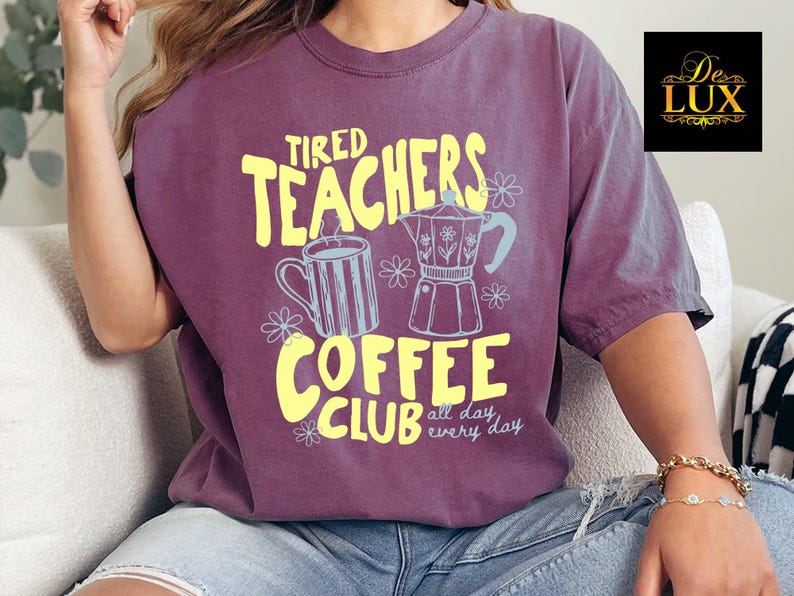 K&ouml;nnte beinhalten: Ein lila T-Shirt mit gelbem Aufdruck "Tired Teachers Coffee Club" mit Illustrationen einer Kaffeetasse und eines Espressokochers. Das Shirt hat eine lockere Passform und kurze &Auml;rmel.