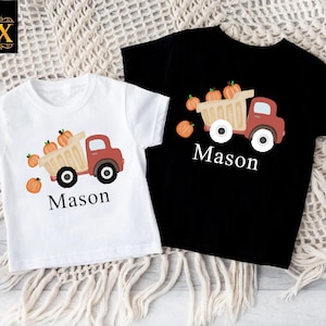 May include: A white t-shirt and a black t-shirt, each featuring a graphic of a red dump truck carrying orange pumpkins. Below the truck, the name Mason is printed in white on the black shirt and in black on the white shirt.