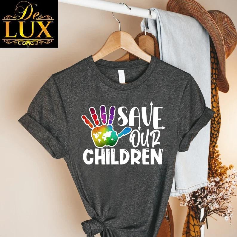 Peut inclure: T-shirt gris fonc&eacute; avec l'inscription blanche "SAVE OUR CHILDREN". Le motif comprend une empreinte de main color&eacute;e avec un globe dans la paume. Le t-shirt est suspendu &agrave; un cintre en bois.