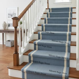 Puede incluir: Una escalera con alfombras azules con una franja color crema en el centro. Las escaleras tienen peldaños y contrahuellas de madera, con una barandilla blanca y un pasamanos de madera. El texto "Nibrant Rugs Store" es visible.