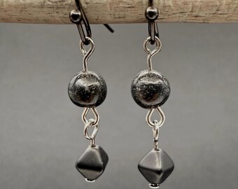 May include: A pair of drop earrings featuring dark metallic hooks, each suspending a round, textured charcoal-coloured bead followed by a smaller, smooth, diamond-shaped black bead connected by silver-toned metal wire loops.