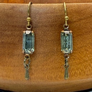 May include: A pair of dangle earrings featuring rectangular, light green faceted stones set in brass-toned metal. Each earring has a hook-style closure and a small, simple metal drop component hanging below the central stone setting.