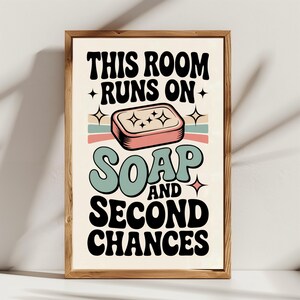 Puede incluir: Un estampado enmarcado con el texto "THIS ROOM RUNS ON SOAP AND SECOND CHANCES". El diseño presenta un estilo retro con una ilustración de jabón y rayas de colores pastel. El marco es de madera.