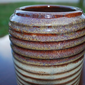 May include: A cylindrical ceramic vase featuring a ribbed texture throughout its body. The upper portion displays a glossy, mottled glaze in shades of deep brown, amber, and gold, while the lower section transitions to a lighter, cream-colored finish.