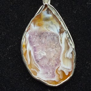 May include: A teardrop-shaped pendant featuring a central cluster of light purple druzy crystals surrounded by bands of translucent white and amber-colored agate. The stone is encased in a polished silver metal setting with a top bail for a chain.