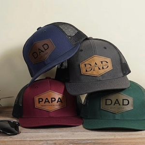 Puede incluir: Un conjunto de gorras de camionero en azul marino, gris carbón, burdeos y verde. Cada gorra tiene un parche de cuero con la palabra "DAD" o "PAPA" y un nombre o fecha. Las gorras tienen una parte trasera de malla y una visera curva.