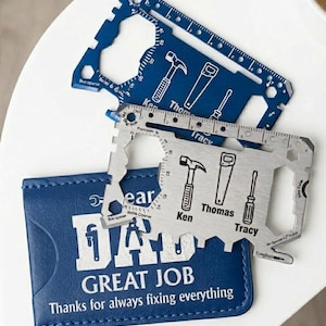 Può includere: Due multiutensili in metallo delle dimensioni di una carta di credito in blu e argento, con icone di martello, sega e cacciavite e i nomi Ken, Thomas e Tracy. Poggiano su un portafoglio in pelle blu con il testo DAD GREAT JOB.