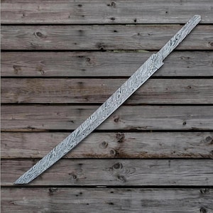 Könnte beinhalten: Eine lange, ungeschliffene Schwertklinge mit einer markanten gemusterten Oberfläche. Die Klinge hat eine gerade Kante und eine Spitze. Das Metall hat ein wirbelndes, marmoriertes Aussehen mit Grau- und Silbertönen. Die Klinge liegt auf einem verwitterten Holzgrund.