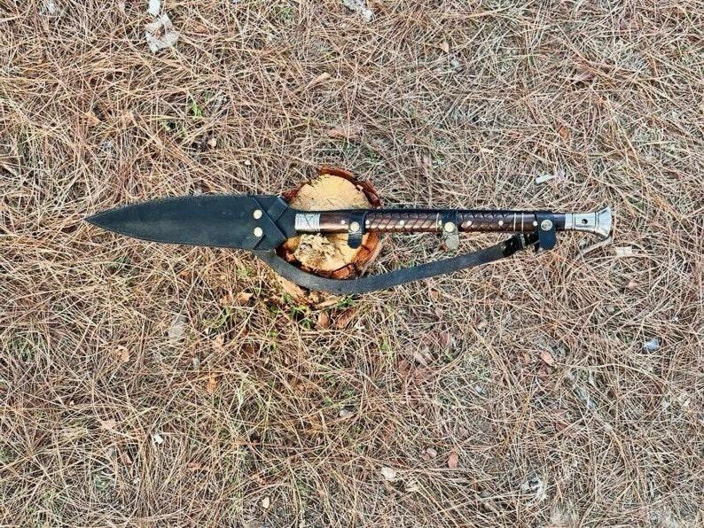 K&ouml;nnte beinhalten: Ein handgefertigter Speer mit schwarzer Klinge und einem mit Leder umwickelten Holzgriff. Der Speer liegt auf einem kleinen Holzsockel und hat einen Lederriemen. Der Speer ist etwa 76 cm lang.