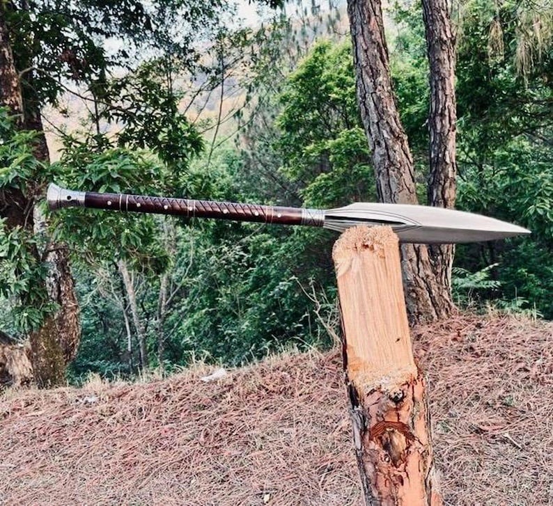 K&ouml;nnte beinhalten: Ein Speer mit einer silbernen Klinge und einem dunkelbraunen Holzgriff. Der Griff hat ein strukturiertes Design und silberne Akzente. Der Speer liegt auf einem Holzpfosten in einer Waldumgebung.