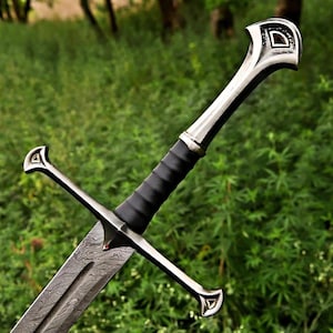 May include: A close-up of a sword with a patterned blade and a black leather-wrapped handle. The sword has a silver hilt with decorative details and a crossguard. The background is a blurred view of green foliage.