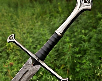 May include: A detailed image of a sword with a patterned blade and a black leather-wrapped handle. The sword features a silver hilt with decorative elements and a crossguard. The background is a blurred view of green foliage.