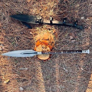K&ouml;nnte beinhalten: Ein Speer mit silberner Klinge und dunklem Holzgriff, der auf einem Baumstumpf ruht. Eine schwarze Lederscheide mit Riemen befindet sich dar&uuml;ber. Die Gegenst&auml;nde liegen auf einem Bett aus trockenen Bl&auml;ttern und Kiefernnadeln.