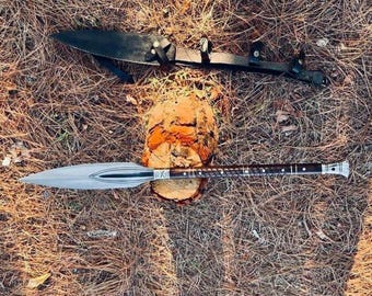 May include: A spear with a silver blade and a dark wooden handle, resting on a tree stump. A black leather sheath with straps is above it. The items are set on a bed of dry leaves and pine needles.