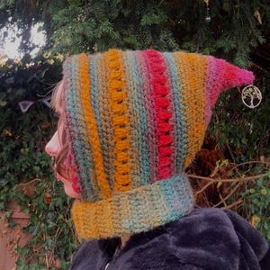 May include: A colorful, handmade crochet hood in shades of yellow, orange, red, and teal. The hood has a pointed tip and a matching neck warmer. The hood is worn by a person.