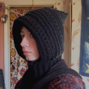 May include: A black, knitted hood with a pointed top. The hood is made of thick yarn and covers the head and neck. The person is wearing a dark jacket underneath the hood.