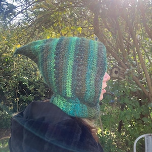 May include: A green and brown striped knitted hood with a pointed tip. The hood is made of yarn and has a cowl neck. The hood is worn over a black coat. The background is a sunny outdoor scene with trees and foliage.