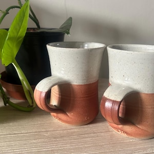 May include: Two handcrafted ceramic mugs, featuring a speckled white upper section and a reddish-orange lower section. The mugs have distinctive handles and are presented on a light-coloured wooden surface. A potted plant is visible in the background.