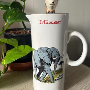 May include: White ceramic pitcher with a handle, featuring a blue elephant illustration and the word "Mixer" in red. A decorative deer head sits on top. The words "African Elephant" are also printed on the pitcher.
