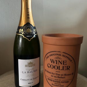 May include: A bottle of Brut champagne with a gold foil top and a terracotta wine cooler. The champagne bottle has a white label with black text. The wine cooler has a circular label with text.