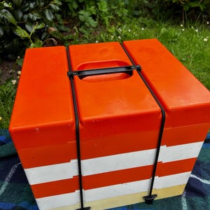 May include: A retro picnic hamper in orange and white stripes, featuring a black handle and straps. The rectangular basket has a hinged lid. It's resting on a blue and green tartan blanket, perfect for outdoor use.