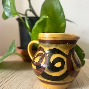 May include: A small, yellow ceramic jug with a handle, decorated with a brown and black spiral pattern. The jug sits on a light wooden surface, with a green plant visible in the background.