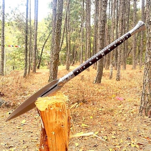 May include: A spear with a long, sharp, silver blade and a dark brown wooden handle. The handle is decorated with silver bands and small white dots. The spear is resting on a wooden stump in a forest setting.