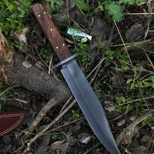 May include: A large, dark-bladed knife with a wooden handle and brass rivets. The blade is a dark gray color, and the handle is a rich brown. The knife is lying on a bed of leaves and twigs, suggesting an outdoor setting.