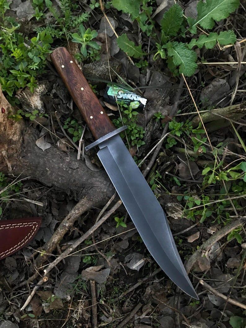 May include: A large, dark-bladed knife with a wooden handle and brass rivets. The blade is a dark gray color, and the handle is a rich brown. The knife is lying on a bed of leaves and twigs, suggesting an outdoor setting.