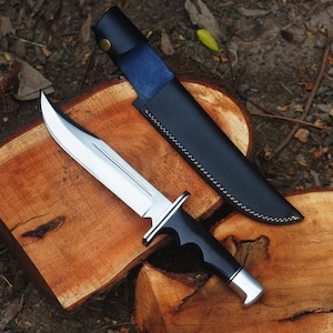 May include: A large hunting knife with a polished steel blade and a black handle, resting on a wooden surface. The knife is accompanied by a black leather sheath with blue accents.