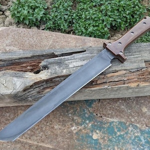 May include: A large, handmade sword with a dark, gray blade and a brown wooden handle. The handle is secured with rivets. The sword is resting on a weathered wooden surface, with greenery in the background.
