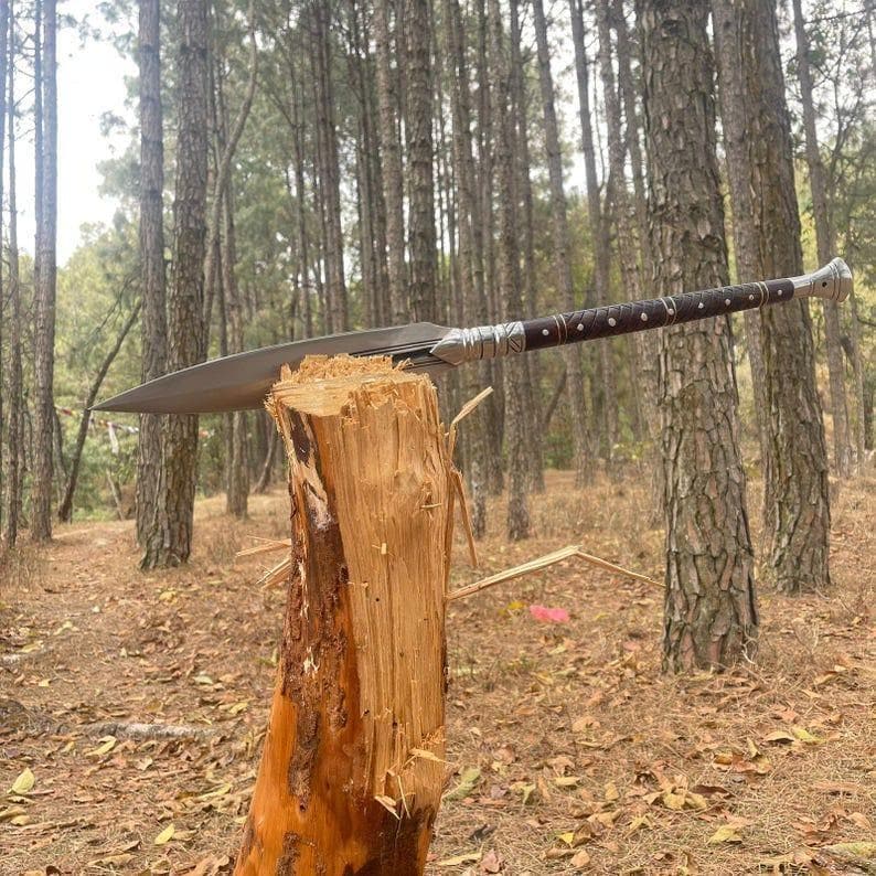 May include: A spear with a long, double-edged blade and a dark brown wooden handle with silver accents. The spear is resting on a chopped tree stump in a forest setting.