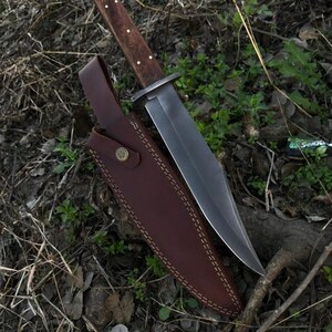 May include: A large, fixed-blade knife with a dark wooden handle and a polished steel blade. The knife rests in a brown leather sheath with decorative stitching and a snap closure. The knife and sheath are displayed on a bed of leaves and twigs.
