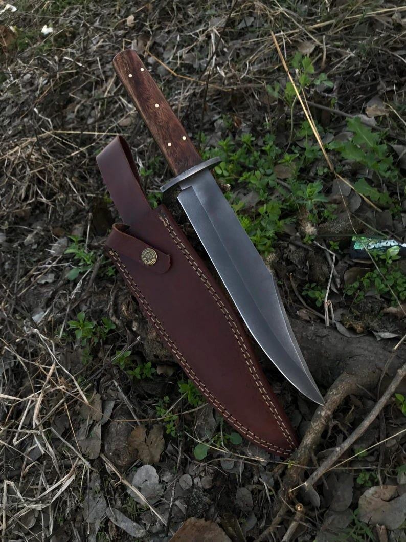 May include: A large, fixed-blade knife with a dark wooden handle and a polished steel blade. The knife rests in a brown leather sheath with decorative stitching and a snap closure. The knife and sheath are displayed on a bed of leaves and twigs.