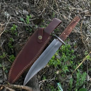 May include: A large hunting knife with a polished steel blade and a wooden handle with brass rivets. The knife is displayed with its brown leather sheath, which has a belt loop and a snap closure. The knife and sheath are resting on a bed of leaves and grass.