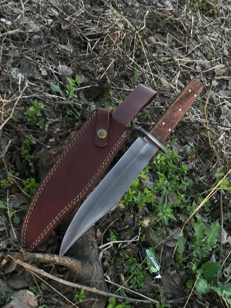 May include: A large hunting knife with a polished steel blade and a wooden handle with brass rivets. The knife is displayed with its brown leather sheath, which has a belt loop and a snap closure. The knife and sheath are resting on a bed of leaves and grass.